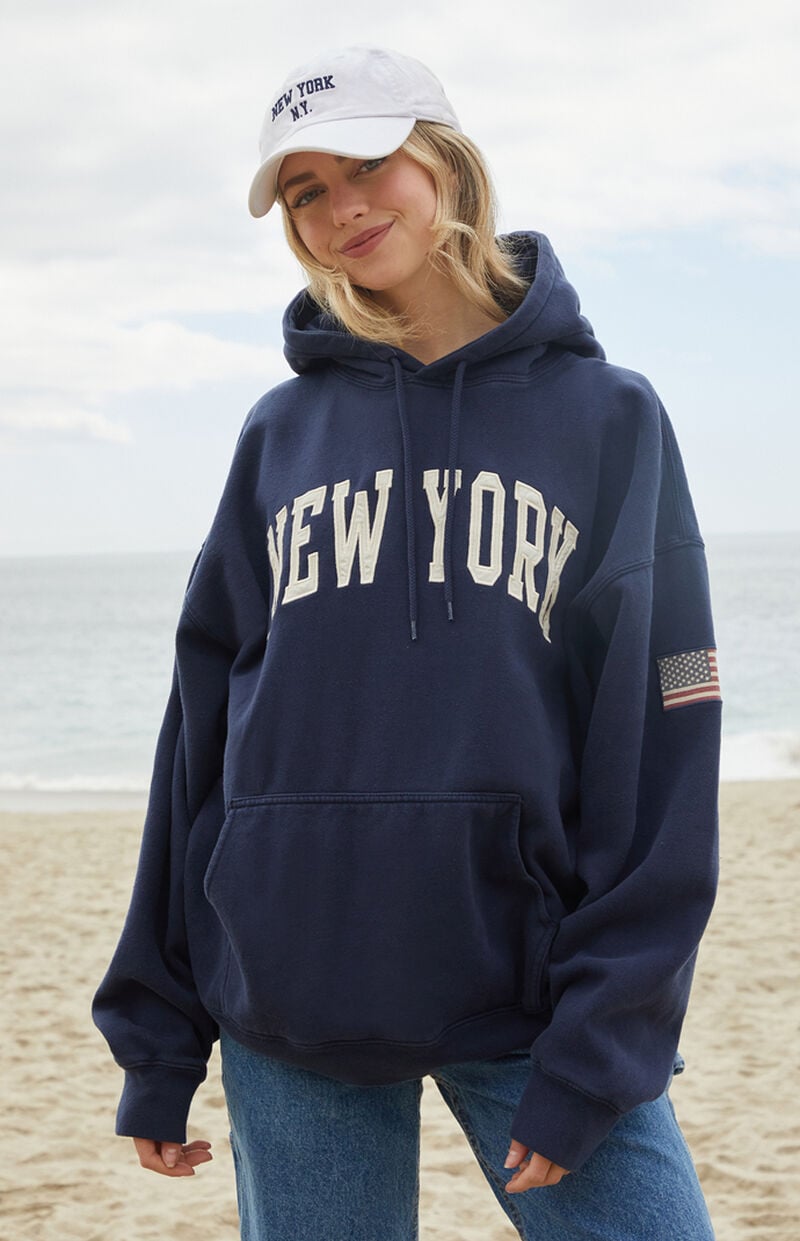 Model at the beach wearing the John Galt Christy New York hoodie with blue jeans and a white cap